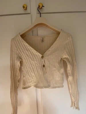 Beige ribbad kofta från My Trend - Snygg beige ribbad kofta från My Trend med två träknappar framtill. Koftan har djup v-ringning, lång ärm och är tillverkad i ett mjukt, stretchigt material. Perfekt att styla med jeans eller kjol för en trendig look.