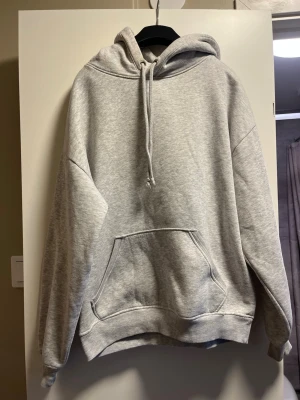 Ljusgrå oversized hoodie med huva - En stilren ljusgrå hoodie i oversized modell med stor känguruficka framtill och justerbar dragsko i huvan. Tillverkad i mjukt sweatshirtmaterial med ribbade muddar vid ärmar och nederkant. Perfekt för en avslappnad och trendig look.