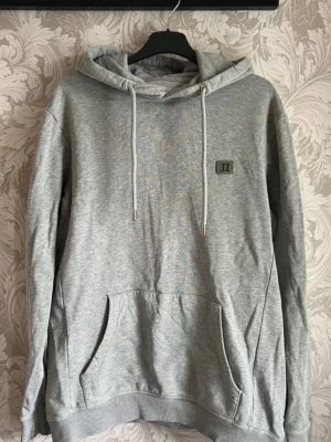 Ljusgrå hoodie från Les Deux - En stilren ljusgrå hoodie med stor magficka och justerbar huva med vita snören. Tröjan har ribbade muddar vid ärmslut och nederkant samt en liten patch med 'II' på bröstet. Perfekt för en avslappnad look och enkel att matcha med andra plagg. Hoodien har en liten skavank vid ett av snörena. Defekten är minimal och produkten säljs i befintligt skick.