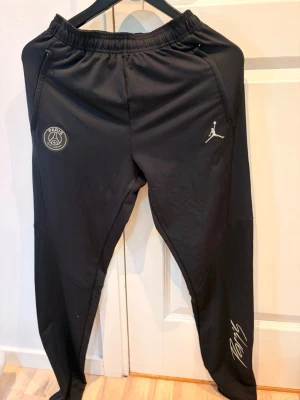 Svarta PSG x Jordan träningsbyxor - Säljer ett par svarta träningsbyxor från PSG x Jordan med elastisk midja, dragkedjefickor och loggor på framsidan. Byxorna har smal passform och är tillverkade i ett lätt, sportigt material. Perfekta för träning eller chill.