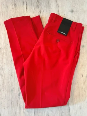 Röda golfbyxor från J.Lindeberg - Snygga röda kostymbyxor från J.Lindeberg med klassisk rak passform och pressveck. Byxorna har bälteshällor, knappstängning och fickor bak. Perfekta för dig som vill sticka ut med färg. Helt oanvända med prislapp. Modellen heter: Ellott pant. Storleken är: 30/32
