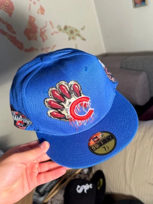 Blå Chicago Cubs keps med brodyr - Säljer en blå fitted keps med broderade detaljer som björn, stadssilhuett och Cubs-logga. Kepsen har platt skärm och coola patches på sidorna, bland annat en Champions-patch. Perfekt för dig som gillar streetwear och sportig stil.