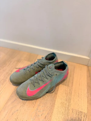 Gråa Nike fotbollsskor med rosa detaljer - Säljer ett par gråa Nike fotbollsskor med snygga rosa detaljer och logga. Skorna har snörning och dobbar för bra grepp på planen. Tillverkade i ett lätt syntetmaterial som ger bra bollkontroll och komfort under matchen.