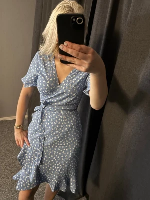 Blå omlottklänning från ONLY - Supersöt blå omlottklänning från ONLY med vitt blommönster. Klänningen har korta ärmar, volangkant nertill och knyt i midjan för en snygg passform. Perfekt för sommarens alla tillfällen!