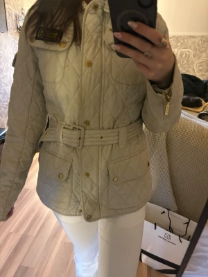 Beige Barbour jacka med bälte - Snygg beige quiltad jacka med bälte i midjan och guldiga knappar. Jackan har fickor framtill, dragkedja och knappar. Dock är det en fläck på jackan vid bröstet (som visas på bilden) perfekt som höst / vårjacka