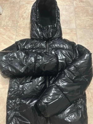 Svart glansig pufferjacka med huva - Säljer en svart, glansig pufferjacka med stor huva och dragkedja framtill. Jackan har en oversized look och är tillverkad i ett blankt syntetmaterial. Perfekt för kalla dagar när du vill hålla dig varm och se trendig ut. Ser helt ny ut🎀. 
