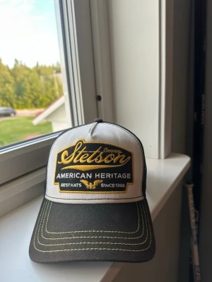 Stetson truckerkeps svart/vit - Snygg truckerkeps från Stetson i svart och vitt med broderad patch framtill där det står 'Stetson American Heritage'. Kepsen har mesh på baksidan för extra ventilation och justerbar snapback. Gula detaljer. Använd under 2-3 veckor 