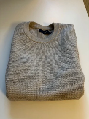 Beige stickad tröja från Riley - Snygg beige stickad tröja från Riley med rund halsringning och diskret strukturmönster. Tröjan har ribbade muddar vid halsen och är tillverkad i ett mjukt material som passar perfekt till lager på lager. Enkel och stilren design.