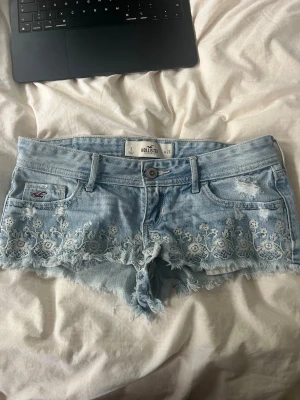 Ljusblå jeansshorts med spetsdetaljer - Supersnygga ljusblå jeansshorts från Hollister med slitna kanter och vita broderade spetsdetaljer längst ner. Klassisk femficksmodell med låg midja och knappgylf. Perfekta för sommaren och ger en chill vibe till din outfit.