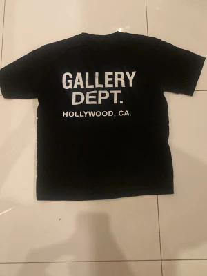 Svart Gallery Dept t-shirt - Säljer en svart t-shirt från Gallery Dept med vit text på både bröstet och ryggen. På ryggen står det 'GALLERY DEPT. HOLLYWOOD, CA.' i stora bokstäver. Klassisk passform och korta ärmar. Perfekt för dig som gillar streetwear och enkel stil.