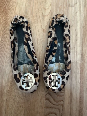 Tory Burch ballerinaskor - Snygga ballerinaskor från Tory Burch i leopardmönstrad mocka med guldfärgad logga framtill. Skorna har rund tå och elastisk kant för skön passform. Innersulan tyvärr borta
