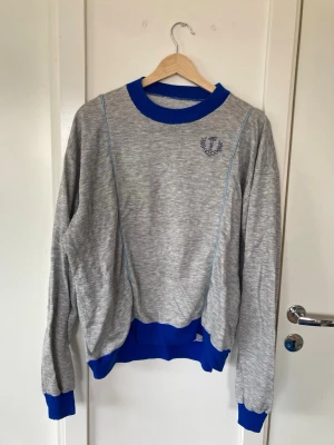 Grå vintage OS tröja  - Snygg grå sweatshirt med blå ribbade muddar, krage och nederkant. Okänt när den är ifrån men OS symbol. Tröjan har fickor och är i storlek L men uppfattar den inte stor i storleken, eventuellt mer som en M 