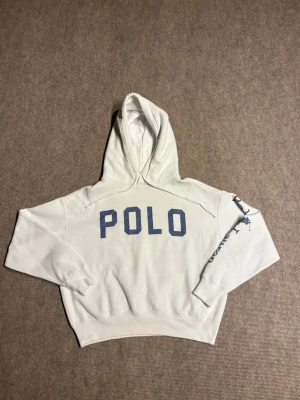 Vit Ralph lauren hoodie med blå POLO-tryck - Säljer en vit hoodie med stor blå POLO-text på bröstet och tryck längs ena ärmen. Hoodien har huva med dragsko och ribbade muddar vid ärmar och midja. Perfekt för dig som gillar en clean och sportig stil.
