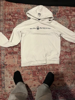 Vit hoodie från Sail Racing - Säljer en vit hoodie från Sail Racing med svart logga och text på bröstet. Hoodien har en stor ficka framtill och klassisk huva. Tillverkad i mjukt bomullsmaterial som är skönt mot huden. Perfekt för en avslappnad och sportig stil.