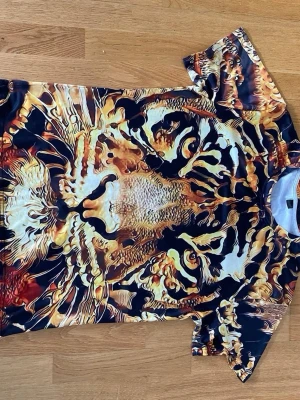 T-shirt med tigertryck i starka färger - Unik t-shirt med ett stort, detaljerat tigeransikte tryckt över hela plagget. Färgerna går i svart, orange, gul och vit och ger ett riktigt iögonfallande intryck. T-shirten har korta ärmar och är tillverkad i ett mjukt, syntetiskt material.
