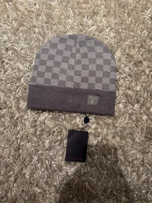 Grå mössa  - Snygg grå mössa  med klassiskt rutmönster och vikt kant. Logga framtill på kanten och medföljande tagg. Perfekt accessoar för dig som vill ha en exklusiv och stilren look.