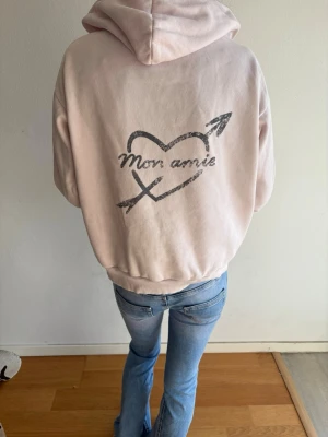 Ljusrosa hoodie med tryck - Ljusrosa hoodie med huva och trycket 'Mon amie' i svart på både bröstet och ryggen. Ryggen har även ett hjärta med pil och korsade pilar. Mjuk och bekväm passform, ribbade muddar vid ärmar och nederkant. Perfekt för en avslappnad look.