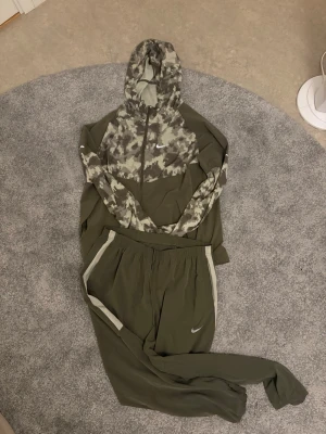 Nike olivgrön loungewear set - Nike loungewear set i olivgrönt med matchande hoodie och byxor. Hoodien har ett digitalt camomönster i grått och grönt samt dragkedja och fickor. Byxorna har elastisk midja och vita detaljer vid fickorna. Perfekt för chill dagar hemma eller när du vill vara bekväm.