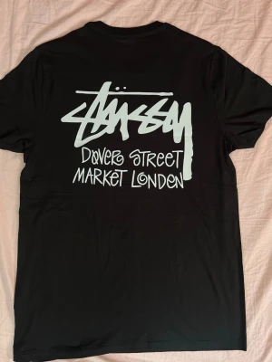 Svart Stüssy Dover Street Market tee - Säljer en svart t-shirt från Stüssy med Dover Street Market London-tryck i vitt både fram och stort på ryggen. Klassisk passform med korta ärmar och rund hals. Perfekt för dig som gillar streetwear och vill sticka ut med en ikonisk logga.