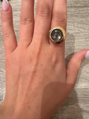 Guldfärgad ring med stor svart sten - Säljer en statementring i guldfärg med en stor, rund svart sten i mitten. Ringen har en bred kant och en modern, iögonfallande design. Perfekt för dig som vill ha något unikt och trendigt på fingret.