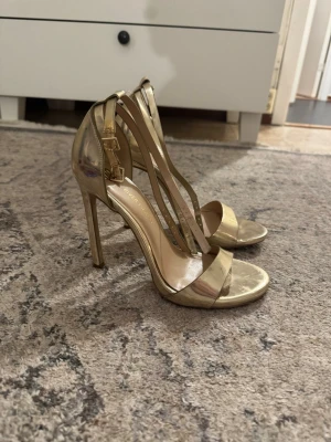 Guldiga höga pumps med remmar - Säljer ett par glansiga guldiga pumps från River Island med smala remmar och öppen tå. Skorna har hög klack och justerbara spännen vid ankeln. Perfekta för att lyfta vilken outfit som helst med sin metallic-finish och eleganta design.