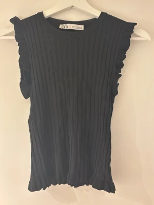 Svart ribbad topp från Zara - Säljer en svart ribbad topp från Zara med volangdetaljer vid axlarna. Toppen är ärmlös och har en rund halsringning. Perfekt att styla med både jeans och kjol för en trendig look.