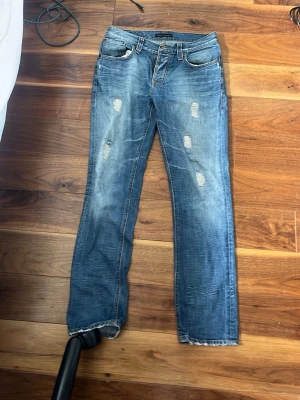 Nudie Grim Trim - Säljer ett par blå jeans med slitningar och raka ben 33/32. Jeansen har klassisk femficksmodell, ljus tvätt och tydliga slitdetaljer framtill och baktill. Perfekta för en avslappnad och trendig look. Säljs pga passar inte mig! Pris kan diskuteras vid snabb affär våga hör av er!