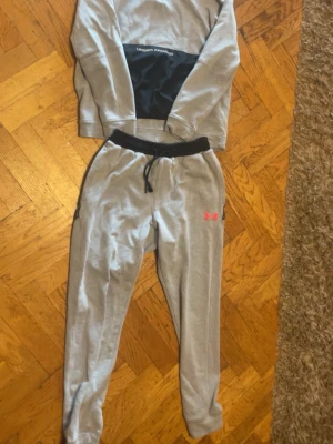 Grått träningsset från Under Armour - Grått träningsset med sweatshirt och byxor från Under Armour. Tröjan har svart panel över magen och byxorna har svart midjeresår med snörning samt rosa logga på benet. Perfekt för sport eller chill.