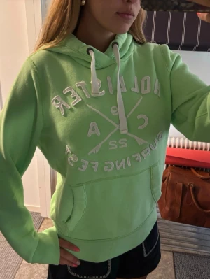 Grön hoodie från Hollister - Snygg ljusgrön hoodie från Hollister med vit broderad logga och text på bröstet. Hoodien har huva med vita dragsnören och en stor magficka. Tillverkad i mjukt bomullsmaterial, perfekt för chill dagar. Klassisk passform och ribbade muddar vid ärmar och nederkant.