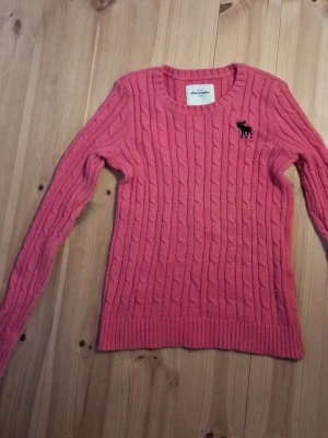 Rosa kabelstickad tröja Abercrombie - Säljer en rosa kabelstickad tröja från Abercrombie med rund halsringning och svart logga på bröstet. Tröjan har långa ärmar och ribbade muddar vid ärmslut och nederkant. Perfekt för dig som gillar klassisk och mysig stil.Tröjan är lite nopprig och har ett par lösa trådar som man ser i sista bilden den är storlek M kids 
