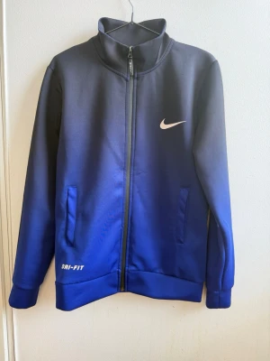 Blå Nike Dri-FIT ziptröja - Snygg ziptröja från Nike i blått med Dri-FIT-material. Tröjan har hög krage, två fickor framtill och vit Nike-logga på bröstet. Perfekt för träning eller chill. Materialet är funktionspolyester som andas och håller dig torr.