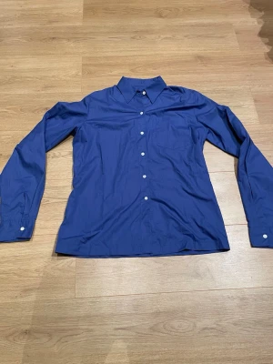 Blå skjorta med vita knappar - Snygg blå skjorta i klassisk modell med långa ärmar och vita knappar framtill. Skjortan har en enkel bröstficka och en stilren krage. Perfekt för dig som gillar en clean och tidlös look.
