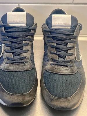 Mörkblå Philippe Model sneakers - Snygga mörkblå sneakers från Philippe Model med detaljer i mocka och textil. Skorna har vita logopatchar på plösen och en ikonisk sköld på sidan. Klassisk låg modell med vita och svarta detaljer på sulan. Perfekta för dig som gillar streetstyle.