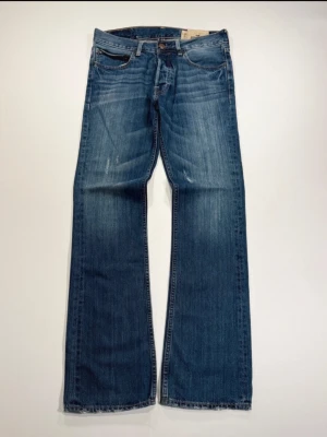 Blå bootcut jeans från Hollister - Säljer ett par blå bootcut jeans från Hollister med klassisk femficksmodell och slitningar framtill. Jeansen har en avslappnad passform och är tillverkade i bomull med snygg tvättad look. Perfekta för en casual och trendig stil.