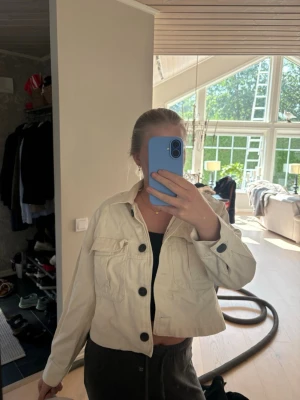 Beige cropped jeansjacka - Snygg beige jeansjacka i croppad modell med stora svarta knappar och två bröstfickor med lock. Jackan har klassisk krage och långa ärmar. Perfekt att slänga över en topp för en avslappnad look.