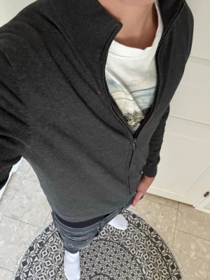 Mörkgrå ziptröja med hög krage - Säljer en stilren mörkgrå ziptröja med hög krage och hel dragkedja framtill. Tröjan har långa ärmar och ribbade muddar vid ärmslut och nederkant. Perfekt att slänga på sig över en t-shirt för en avslappnad look.