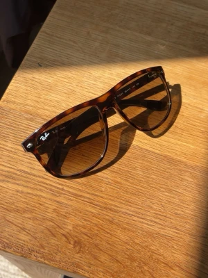 Bruna Wayfarer solglasögon Ray-Ban - Säljer ett par klassiska solglasögon från Ray-Ban i brunmelerad färg. Snygg tidlös design med Ray-Ban-logga på bågen. Tillverkade i Italien och perfekta för soliga dagar.