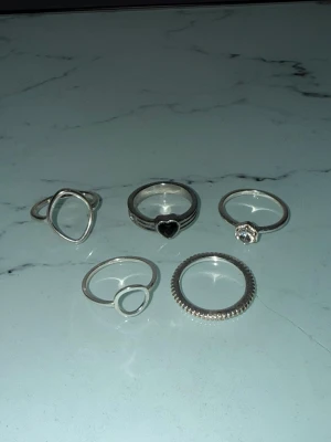 Fem silverfärgade ringar i olika stilar - Säljer ett set med fem silverfärgade ringar i olika designer. En ring har ett svart hjärta, en har en klar sten, två har öppna cirkeldetaljer och en har en räfflad kant. Perfekta för att mixa och matcha till din egen stil. Ringarna är i storlek L, och är inte i äkta silver.