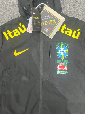 Svart Brasil Nike vindjacka med huva - Snygg svart vindjacka från Nike med Brasil-logga och sponsormärken i gult och grönt. Jackan har huva, dragkedja framtill och justerbara ärmslut. Tillverkad i GORE-TEX-material som står emot regn och vind. Perfekt för dig som gillar sportig stil.