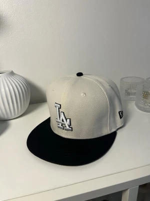 Vit och svart LA-keps från New Era - Snygg vit keps med svart skärm från New Era, prydd med LA-logga framtill och broderad text 'Los Angeles Dodgers' baktill. Klassisk rak skärm och broderade ventilationshål. Perfekt för dig som gillar streetwear och sportig stil.