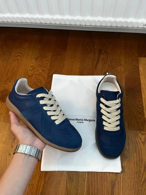 Blå Maison Margiela sneakers - Snygga blå sneakers från Maison Martin Margiela med beige snörning och klassisk gummisula. Skorna har en låg silhuett och är tillverkade i en mix av mocka och läder för en stilren look. Perfekta för dig som gillar minimalistisk design.