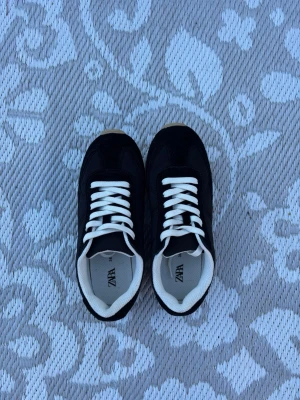 Svarta sneakers från ZARA - Stilrena svarta sneakers från ZARA med vita snören och vit sula. Skorna har en klassisk låg profil och är tillverkade i en mix av textil och syntetmaterial. Perfekta för en trendig och avslappnad look.