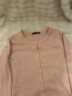 Ljusrosa kofta från 157 - Söt ljusrosa kofta från 157 med rund halsringning och matchande knappar framtill. Koftan är stickad i ett mjukt material och har långa ärmar. Perfekt att slänga över en topp för en chill look.