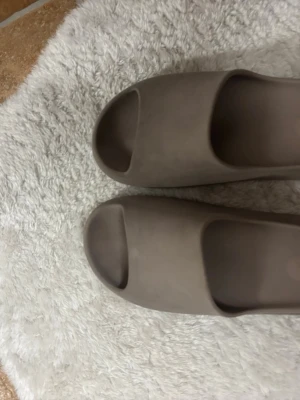 Yeezys slides Beige/brun  - Säljer ett par beige yeezys slides  tofflor med öppen tå och bred rem. Tillverkade i mjukt gummiliknande material för extra komfort. Sulan är räfflad för bättre grepp och tofflorna har en minimalistisk design som passar till många stilar.