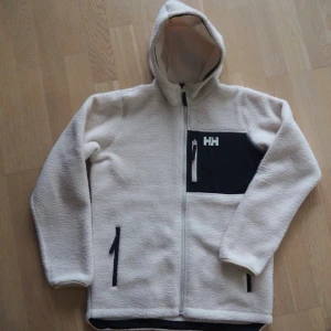 Vit teddyjacka med huva från Helly Hansen - Mysig vit teddyjacka med huva från Helly Hansen. Jackan har hel dragkedja, svart bröstficka med HH-logga och två svarta sidofickor. Tillverkad i fluffigt teddy-material som håller dig varm och stilen på topp. Storlek 164 men passar upp mot 170.                 Sonen fick i födelsedagspresent men ångrade sig när det var för sent att lämna tillbaka. Så nu utan lappar, endast testad. 