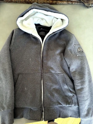 Grå pälsfodrad hoodie Moose Knuckles - Mysig grå hoodie från Moose Knuckles med vitt pälsfoder i huvan och dragkedja framtill. Jackan har två stora fickor, ribbade muddar och en ikonisk logga i metall på ärmen. Perfekt för kalla vinterdagar och riktigt snygg streetstil.