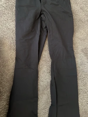 Gråa kostymbyxor med fickdetaljer - Snygga gråa kostymbyxor med raka ben och två fickor med lock bak. Byxorna har bred linning med knappdetaljer och är tillverkade i ett slätt tyg som ger en stilren look. Perfekta för dig som gillar klassisk men modern stil.