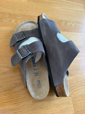 Bruna Birkenstock Arizona sandaler - Snygga bruna Birkenstock Arizona sandaler i mocka med två justerbara remmar och metallspännen. Fotbädden är i mjukt läder och sulan är i kork och gummi för extra komfort. Perfekta för en avslappnad och trendig stil.