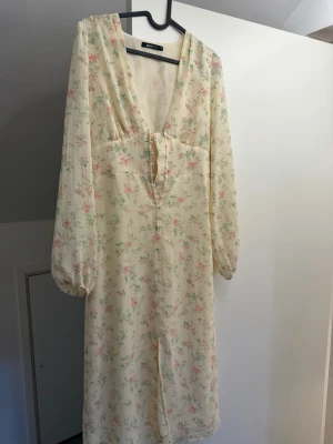 Ljus blommig långklänning med v-ringning - Romantisk långklänning i ljus färg med blommigt mönster i rosa och grönt. Djup v-ringning, knappar framtill och långa ärmar med lätt puff. Perfekt för sommarens alla tillfällen. Inte lång kan skicka fler bilder 