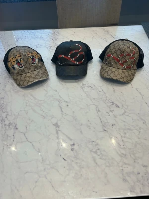 Gucci kepsar med orm och tiger - 500 STYCK! 1500 FÖR ALLA! Tre snygga Gucci kepsar med klassiskt GG-mönster. En keps har en tigerdesign, de andra två har en röd-vit orm. Alla har svart mesh baktill och justerbart band med gröna och röda detaljer. Perfekt för dig som vill sticka ut med lyxig streetstyle.